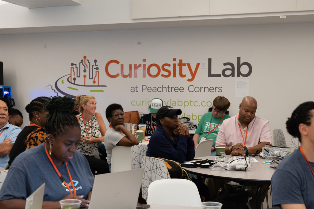Educators sit around tables in front of a wall emblazoned with Curiosity Lab at Peachtree Corners and a circuit diagram logo.
