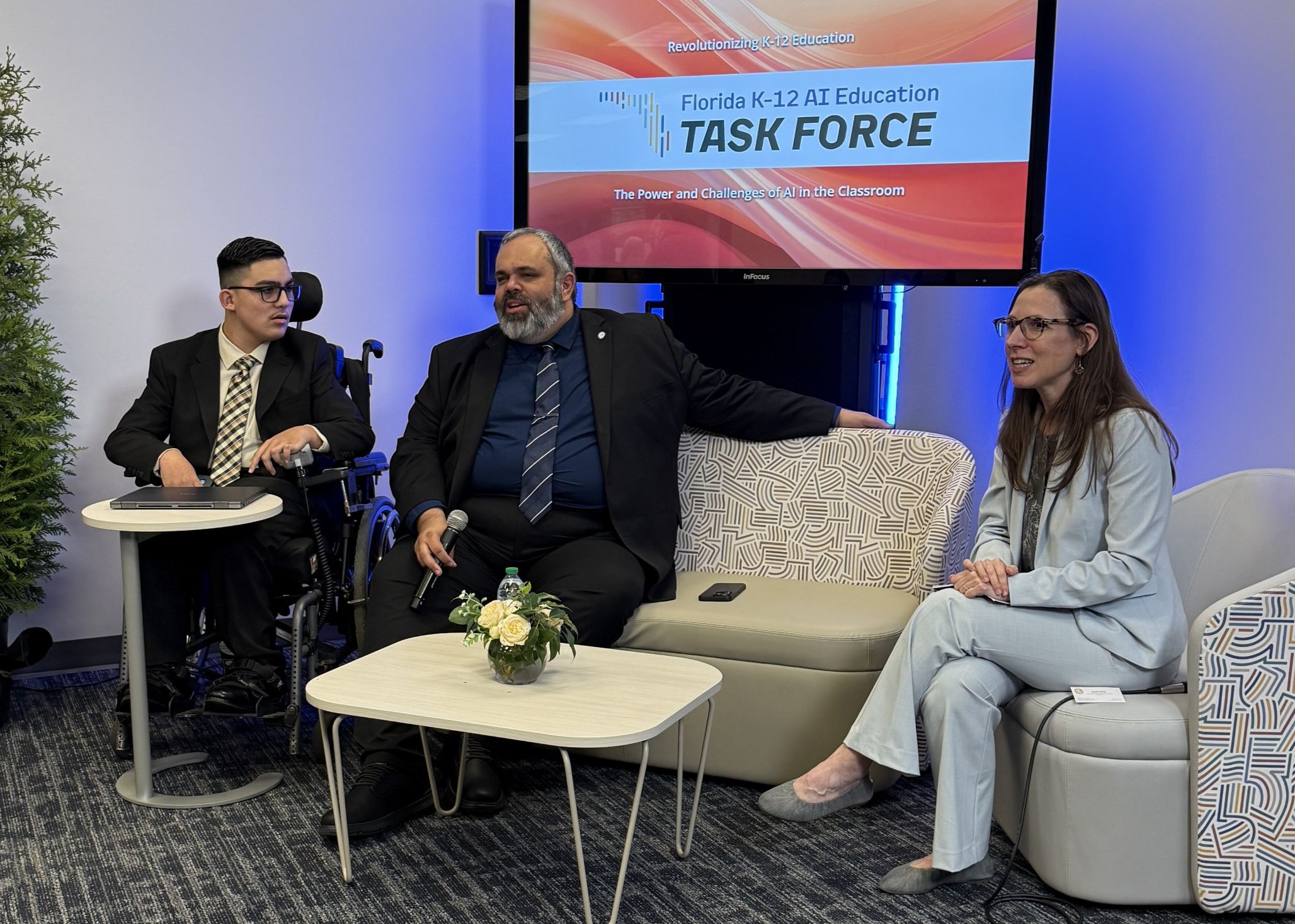 Panel discussion scene: three people in business attire are seated—left participant in a wheelchair, center speaker holding a microphone on a patterned sofa, and right participant in a light-blue suit—around a small table with flowers and a water bottle; behind them a screen reads “Florida K‑12 AI Education Task Force: The Power and Challenges of AI in the Classroom.”